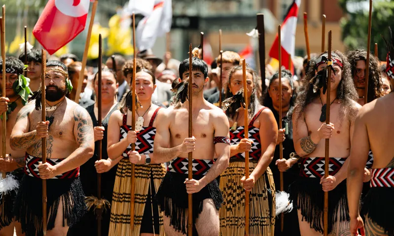 A group of Māori men and women dressed in traditional clothing stand together holding wooden spears during a cultural parade or ceremony, with flags and a crowd visible in the background.