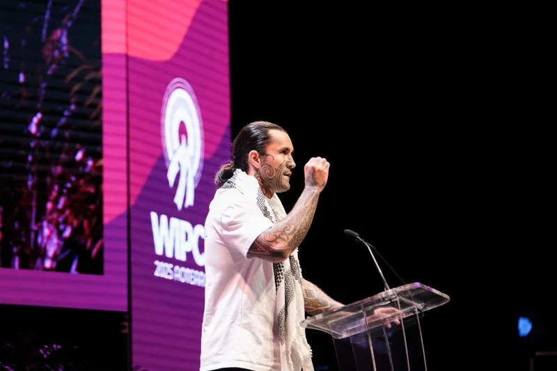 A tattooed man with long hair tied back speaks passionately at a clear podium on stage, with a large purple and pink WIPC 2023 conference screen in the background.