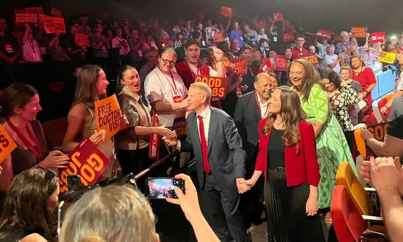 Chris Hipkins, hand in hand with fiancee Toni, walks through a crowd of Labour supporters while reaching out to shake a member's hand. Behind him is Willie Jackson, Carmel Sepuloni and the rest of the Labour caucus.