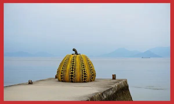 A large yellow pumpkin sculpture with black dots sits at the end of a concrete pier, overlooking calm water with distant blue mountains and a boat in the background. The image has a red border.