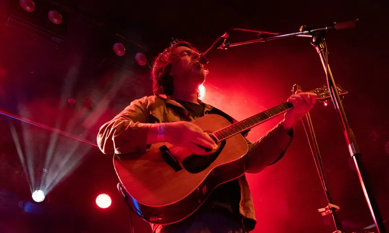 A man playing guitar and singing into a microphone. The room is red with lighting.