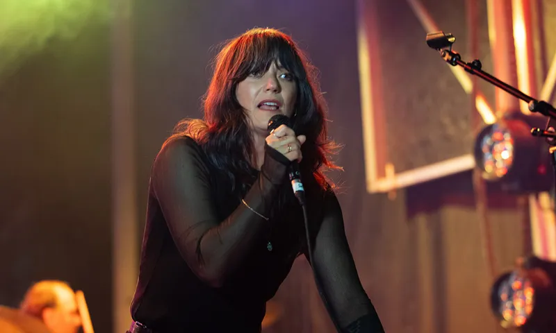 A woman with long black hair and red lipstick singing into a microphone.