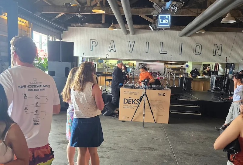 People gather indoors near a small stage with musical instruments at the Pavilion. A person stands at a microphone while another works nearby; some attendees watch, and the space has an industrial, open-ceiling design.