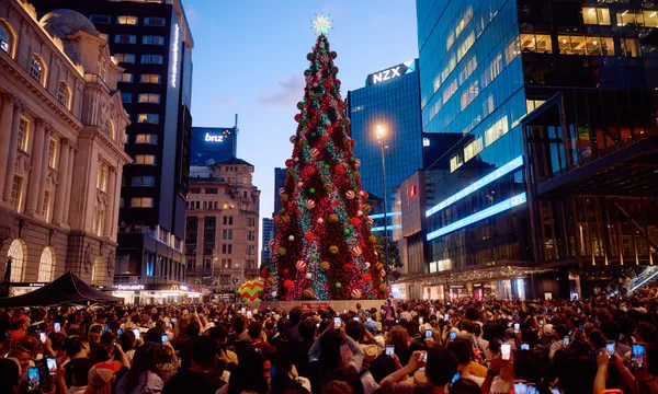 A large crowd gathers around a tall, brightly decorated Christmas tree in an urban plaza at dusk, surrounded by modern glass buildings and illuminated signs. Many people are taking photos with their phones.