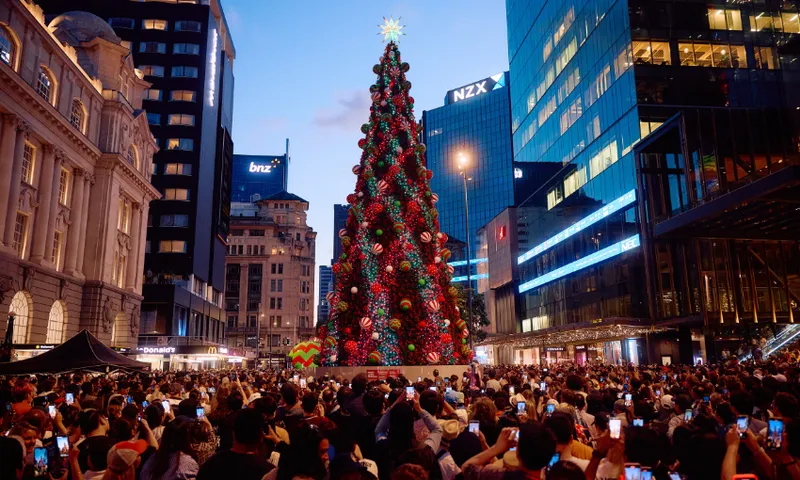 A large crowd gathers around a tall, brightly decorated Christmas tree in an urban plaza at dusk, surrounded by modern glass buildings and illuminated signs. Many people are taking photos with their phones.