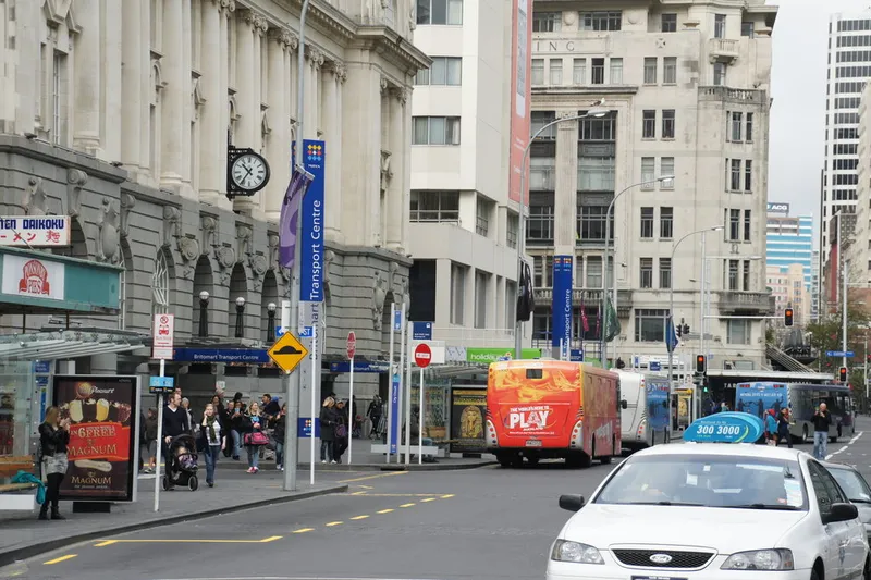 A busy city street scene with people at a bus stop, a red bus, a blue taxi, and tall buildings in the background. There are street signs, a clock, and storefronts visible along the sidewalk.