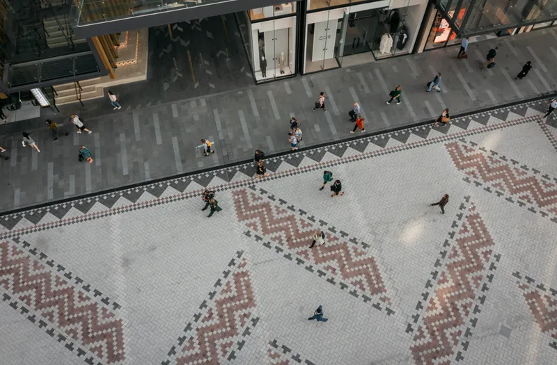 Aerial view of people walking across a patterned plaza with geometric red and white tiles, adjacent to a gray walkway and storefronts with glass windows.
