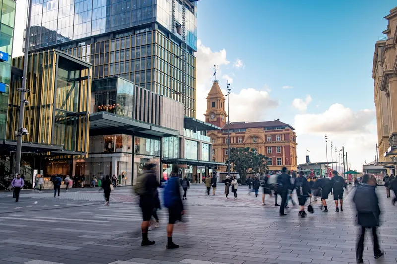 People walk across a modern city square with glass buildings and a historic clock tower in the background under a blue sky with clouds.