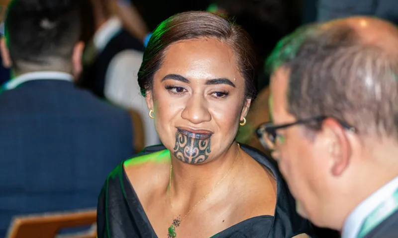A woman with a traditional Māori chin tattoo, wearing a black dress and green necklace, sits beside a man in glasses at a formal event with other attendees in the background.