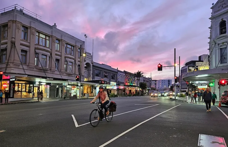 A person rides a bicycle through a city intersection at dusk, passing shops and restaurants with glowing signs under a pink and purple sky. Cars and pedestrians are visible in the background, and traffic lights are red.