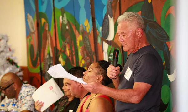 A man with gray hair speaks into a microphone while holding papers, seated beside others at a table. A colorful mural with birds and trees is in the background.
