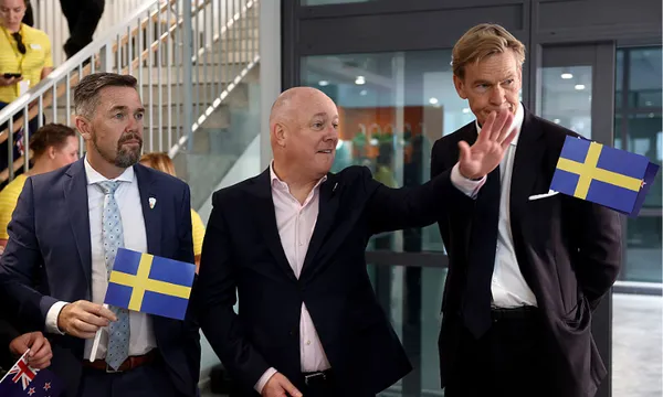 Three men in suits stand indoors, two holding small Swedish flags. One man in the center waves, while others look in different directions. People and a staircase are visible in the background.