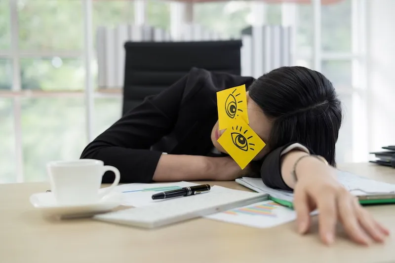 A person in business attire naps at a desk with their head resting on papers, wearing sticky notes with cartoon eyes drawn on them over their closed eyes. A coffee cup, pen, and keyboard are nearby.