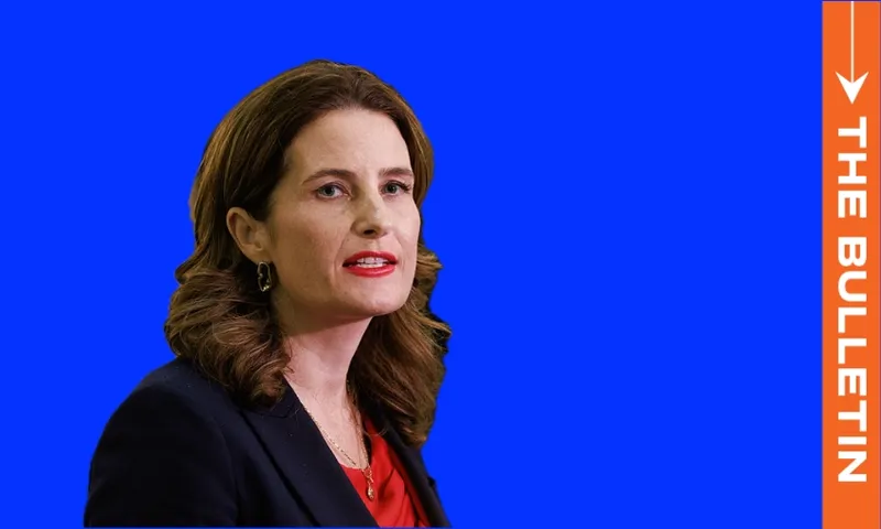 A woman with wavy brown hair in a navy blazer and red top speaks in front of a bright blue background, next to a vertical orange banner reading “THE BULLETIN.”.