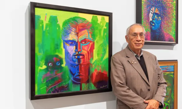 An older man in a brown suit stands beside a colorful, abstract portrait featuring a face with vibrant red, blue, and green hues, displayed in an art gallery. More colorful artwork is visible on the wall behind him.