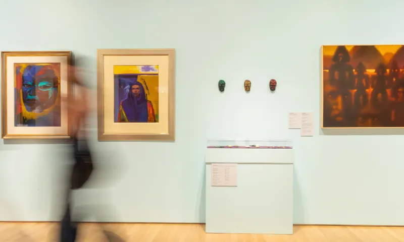 Blurred figure walks past an art gallery wall displaying two colorful portraits, three small sculpted masks, and a glass case with small objects beneath them. The wall is painted light blue.