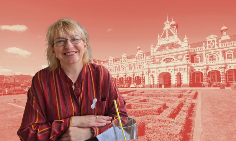 Sophie Barker wears a red dress and smiles at the camera. IN the background there is a photo of the Dunedin railway station