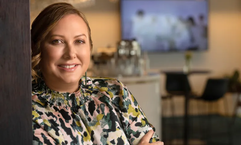 A woman with light brown hair and a patterned blouse smiles while leaning against a wooden surface in a modern, softly lit office or cafe setting. The background is blurred, showing tables and chairs.