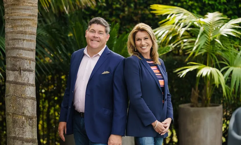 A man wearing a blue blazer stands baack to back with a woman wearing a blue blazer. They are both smiling at the camera