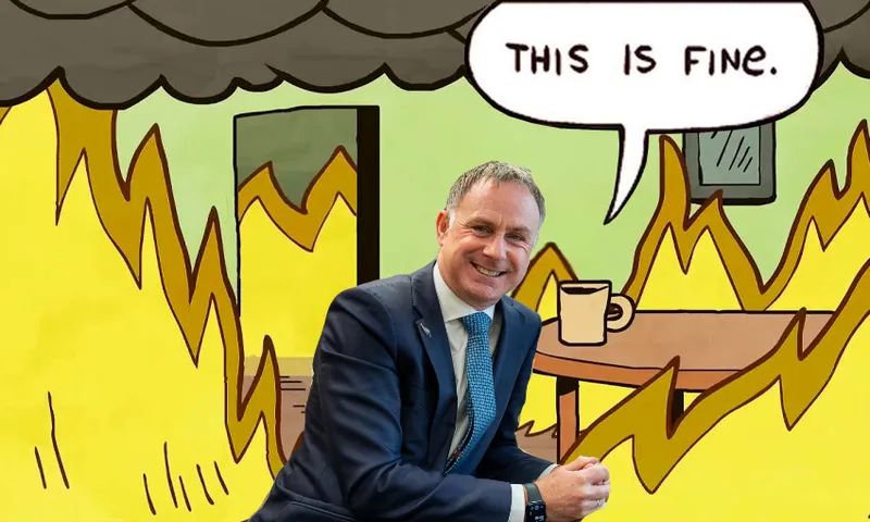 A man in a suit is smiling and sitting in the foreground of the popular "This is fine" meme, which shows a room on fire with a speech bubble saying "This is fine.