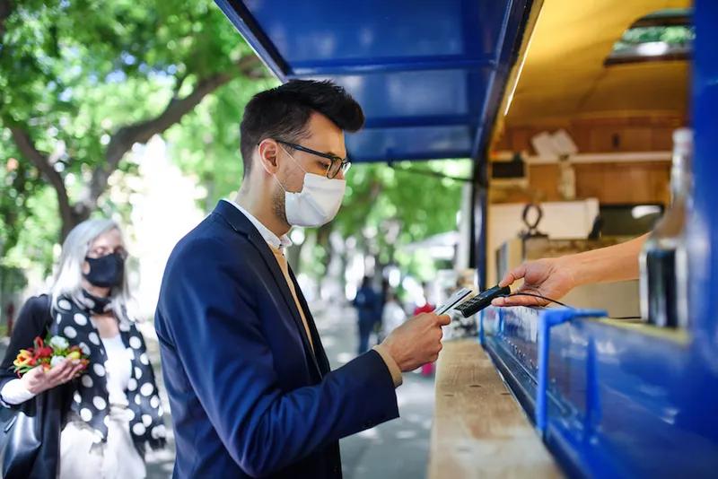 Portrait of young businessman with face mask buying coffee outdoors in city.