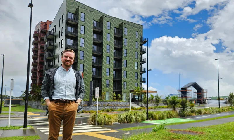 A smiling man stands on a sidewalk in front of a modern, multi-story apartment building on a bright, partly cloudy day. Lush greenery and a crosswalk are visible in the foreground.