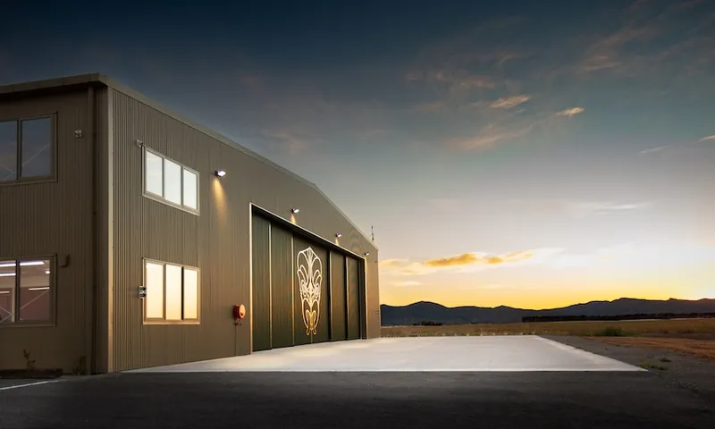 A beige metal hangar with large sliding doors featuring an intricate design, illuminated by exterior lights at dusk, with mountains and an open sky in the background.