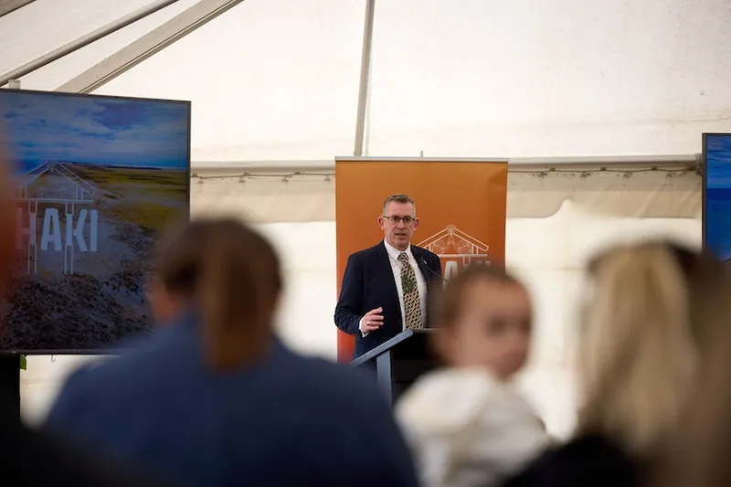 A man in a suit speaks at a podium inside a tent, with an orange backdrop and TV screens on either side. Blurred audience members are visible in the foreground, including a small child.