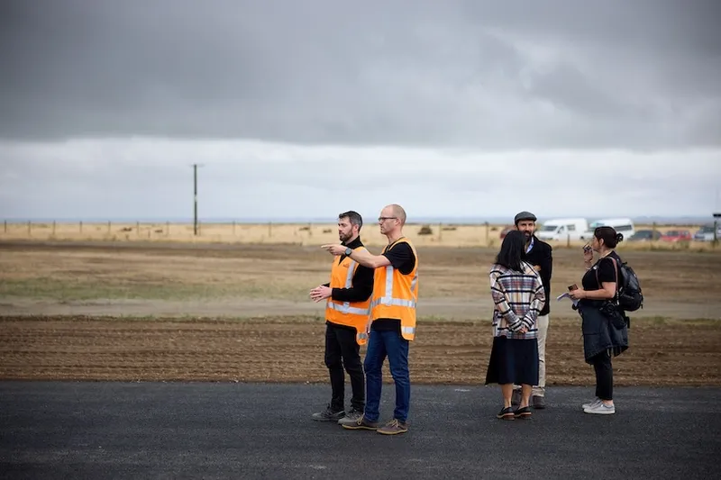 Several people stand outdoors under a cloudy sky, two wearing orange safety vests and gesturing. The group is on a paved area near a field with dry grass in the background.