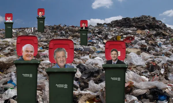 Three green Auckland Council wheelie bins and several in the background are placed in a landfill, each with a cutout photo of a man’s head inside the open lids. Trash piles and a partly cloudy sky are visible.