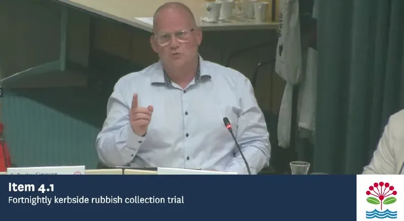 A man in a white shirt and glasses speaks, gesturing with his right hand, at a meeting table. A sign at the bottom reads "Item 4.1 Fortnightly kerbside rubbish collection trial.