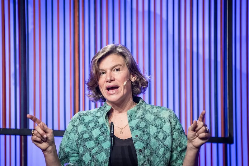 A woman with short, light brown hair speaks on stage, wearing a green patterned shirt and a headset microphone, gesturing with both hands in front of a background with vertical blue and orange lines.