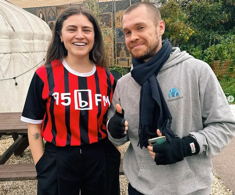 Two people smiling outdoors. The person on the left wears a red and black striped 95bFM shirt; the person on the right wears a grey hoodie, scarf, and fingerless gloves, holding a phone and giving a thumbs up. There’s greenery in the background.