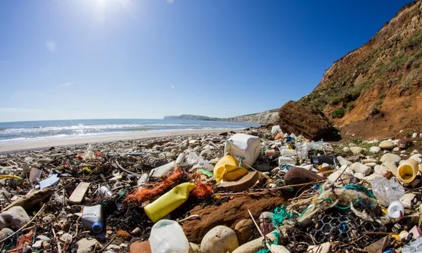 A rocky beach covered with plastic waste and debris under a clear blue sky, with cliffs on one side and the ocean in the background. Sunlight shines brightly over the polluted shoreline.