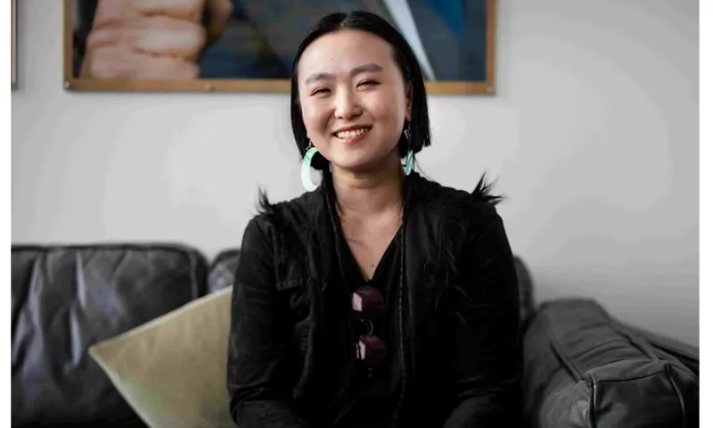 A photograph of poet Jiaqiao Liu, who is a young Asian woman wearing black and sitting on a black leather couch. She is similing.