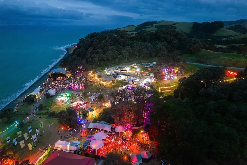 Aerial view of a vibrant outdoor festival at dusk near the coastline, with colorful lights, tents, crowds, and forested hills surrounding the site, beside the ocean.