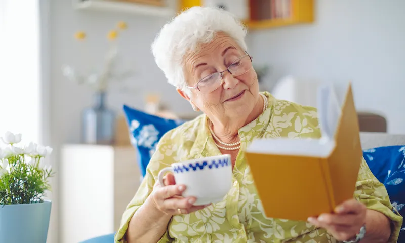 Senior woman holding a cup of tea and a book, looking delighted
