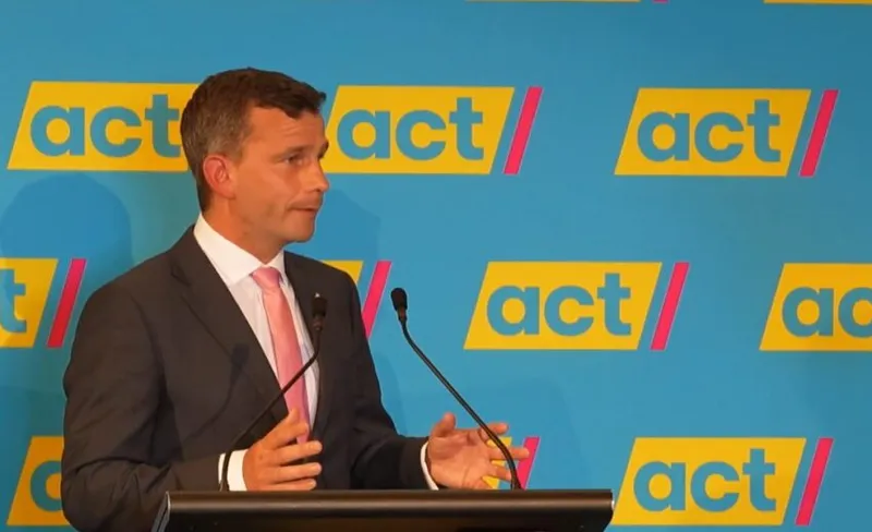 David seymour, a light-skinned man in a grey suit with a pink tie stands in front of a microphoone and the blue, yellow, and pink ACT logo