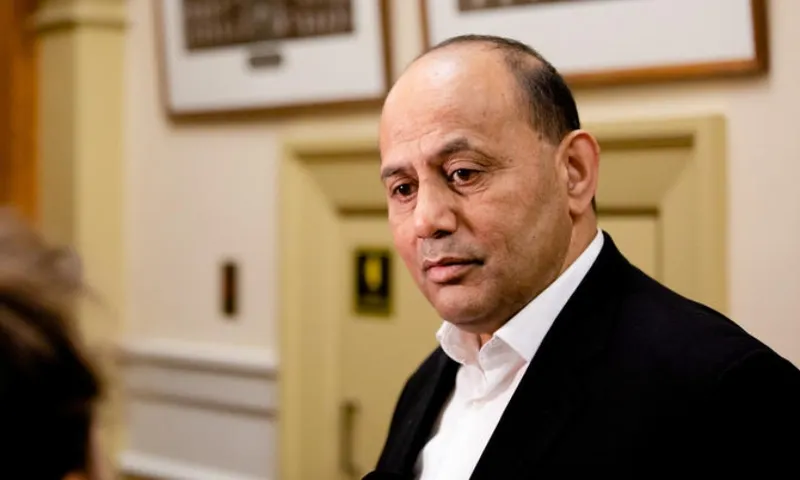 A man in a suit stands indoors, looking thoughtful while speaking to someone. Framed pictures hang on the wall behind him.