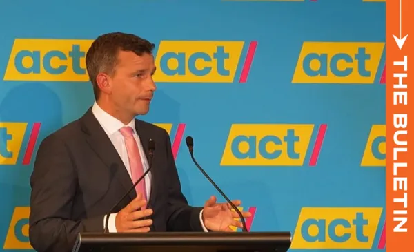 Man in a suit stands at a podium speaking into microphones. The background displays "act" repeated multiple times. An orange vertical banner on the right reads "THE BULLETIN.