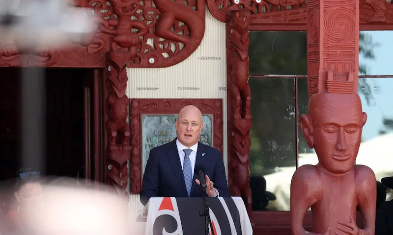 A man in a suit speaks at a podium in front of an ornately carved Māori meeting house, with detailed red wood carvings and a large carved figure visible behind him.