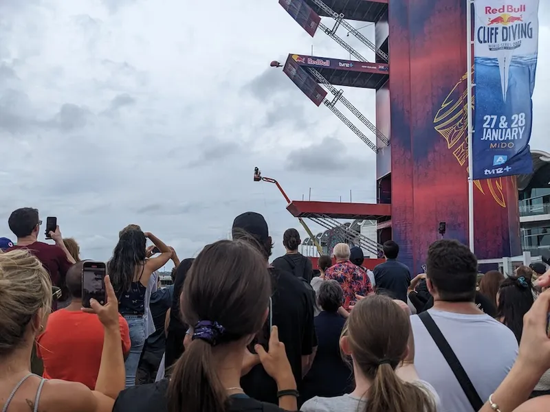 a whole lot of people watching someone tuck as they jomp off a pretty tall platform