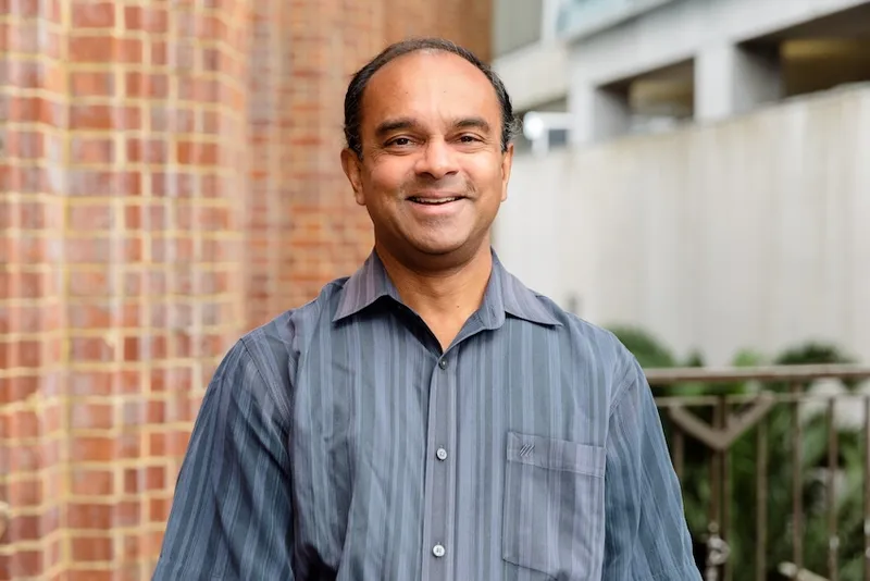 Gehan, a brown skinned man with a slightly receding hairline and a a friendly smile grins in front of a brick wall