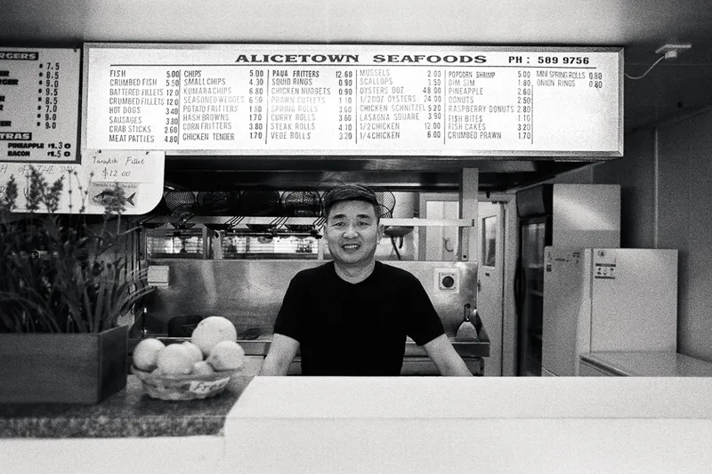 A black and white photo of a man behind the counter at his fish and chip shop with menu above and behind him