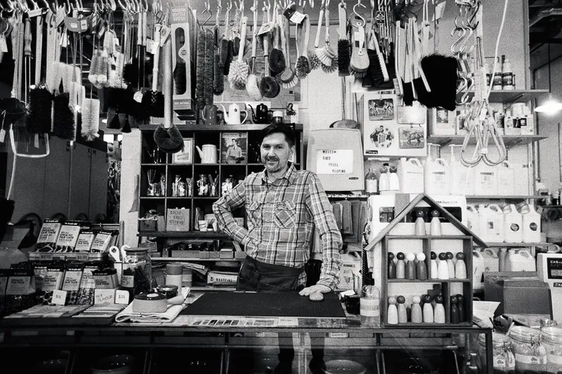 A man stands in his household cleaning store, with various scrubbing brushes hanging from the ceiling above him