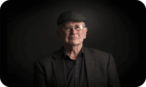 Photographic portrait of an older man in a dark tweed jacket and cheesecutter hat. Beautifully lit so that only his face is really visible. An air of great warmth and cleverness.