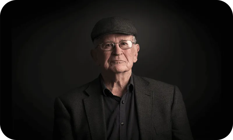 Photographic portrait of an older man in a dark tweed jacket and cheesecutter hat. Beautifully lit so that only his face is really visible. An air of great warmth and cleverness.