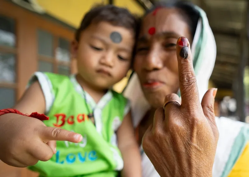 a woman with medium brown skin, wearing a scarf over her head and holding a child with a green shirt, with a finger staned with dark brown/red ink in the foreground