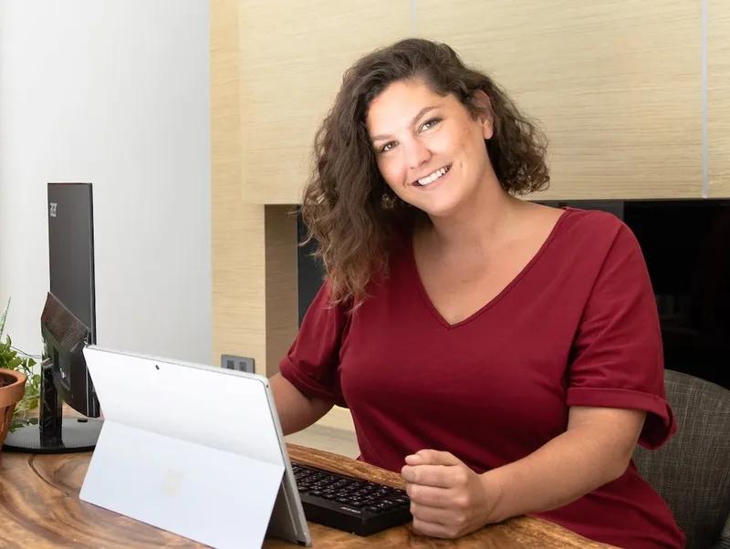 a light skinned woman with medium brown curly hair sitting in front of a folding tablet and smiling