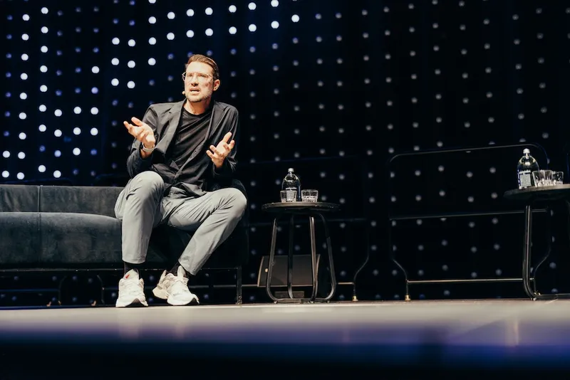 a slightly grizzled white man with glasses and jeans on a couch with a snazzy looking coferency background. he's talking